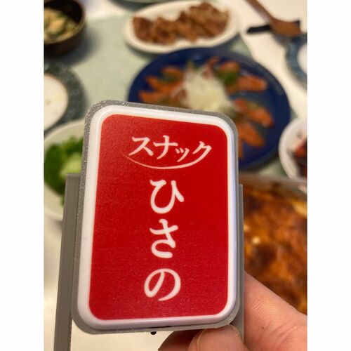食卓を前に指でつまめるサイズの「スナックひさの」看板を持った写真
